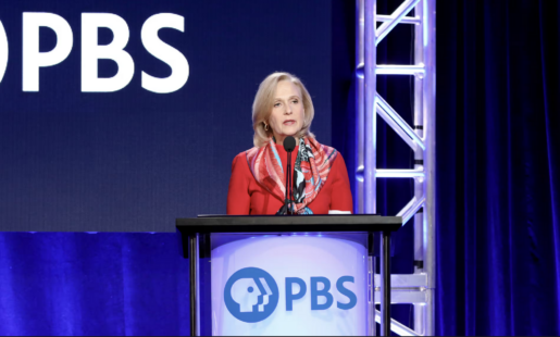 Person wearing red standing and speaking from behind a podium that says PBS.